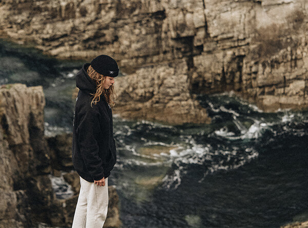 Jeune femme en hoodie et bonnet Studio Quorpo, debout au bord d'une falaise, contemplant l'océan déchaîné, symbolisant l'esprit d'aventure et de liberté.