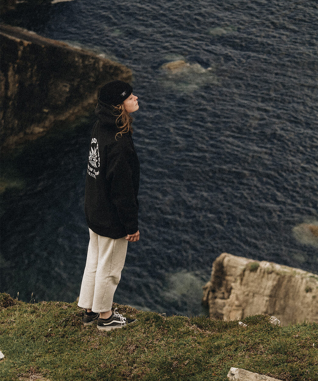 Marie Dromard portant un hoodie et un bonnet Studio Quorpo, debout au bord d'une falaise surplombant l'océan, regardant au loin.