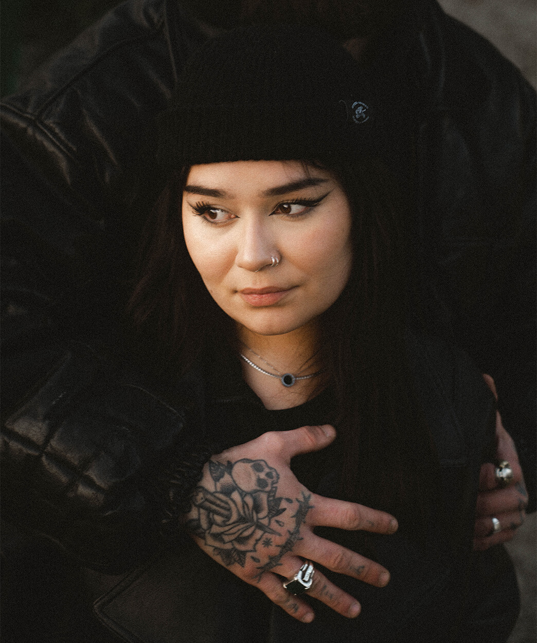 Jeune femme portant un bonnet noir Studio Quorpo, entourée par des bras tatoués, avec un regard pensif et complice.