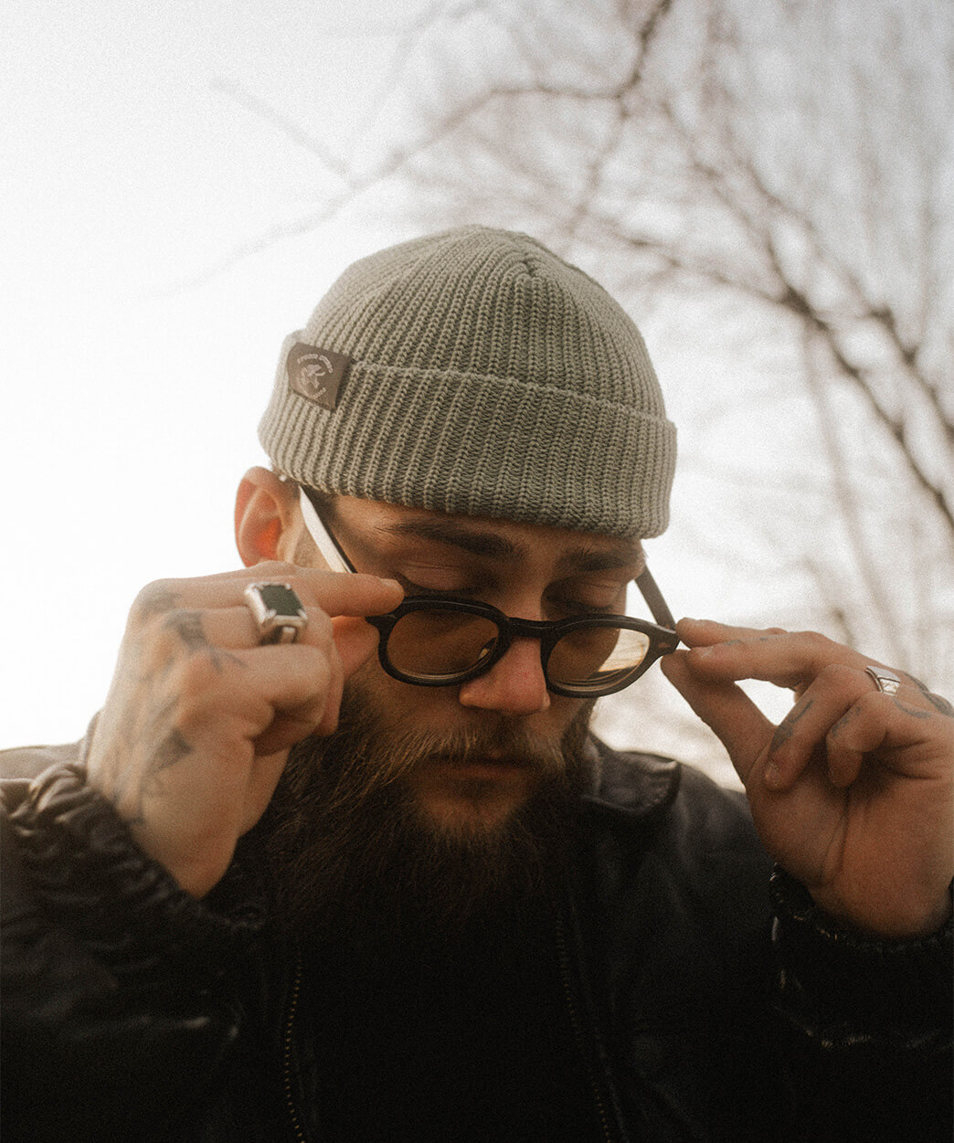 Homme portant un bonnet vert Studio Quorpo, ajustant ses lunettes, avec un regard concentré et un style décontracté, en extérieur.