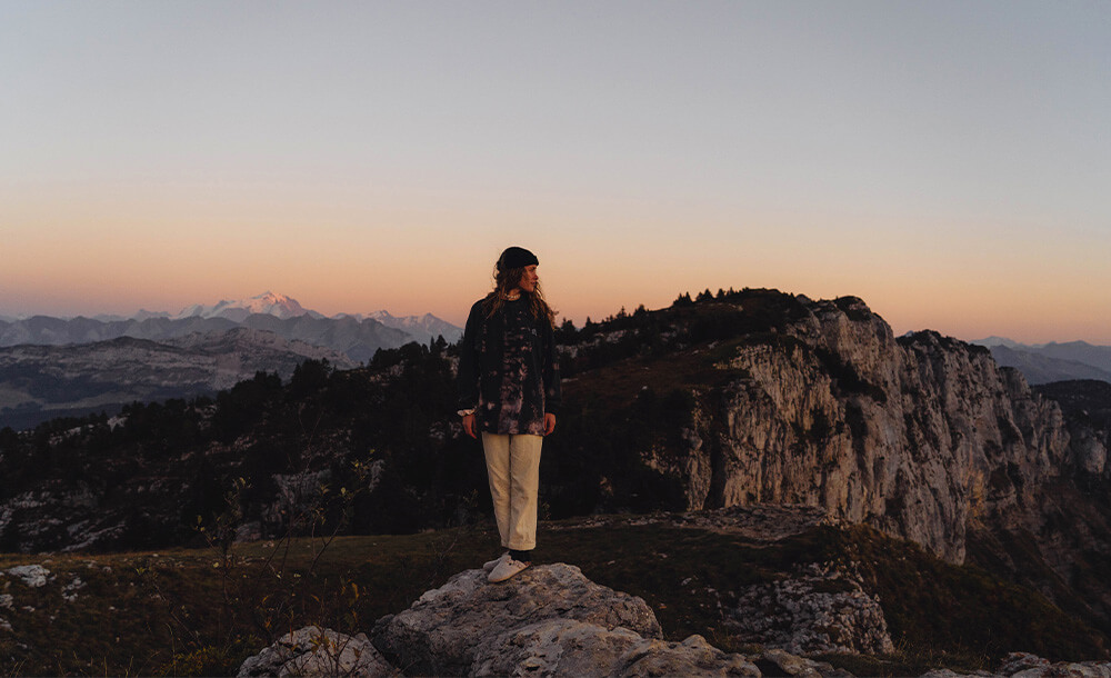 Jeune femme portant un tee shirt à manches longues tie dye Studio Quorpo, debout sur une falaise au coucher de soleil, avec des montagnes en arrière-plan.