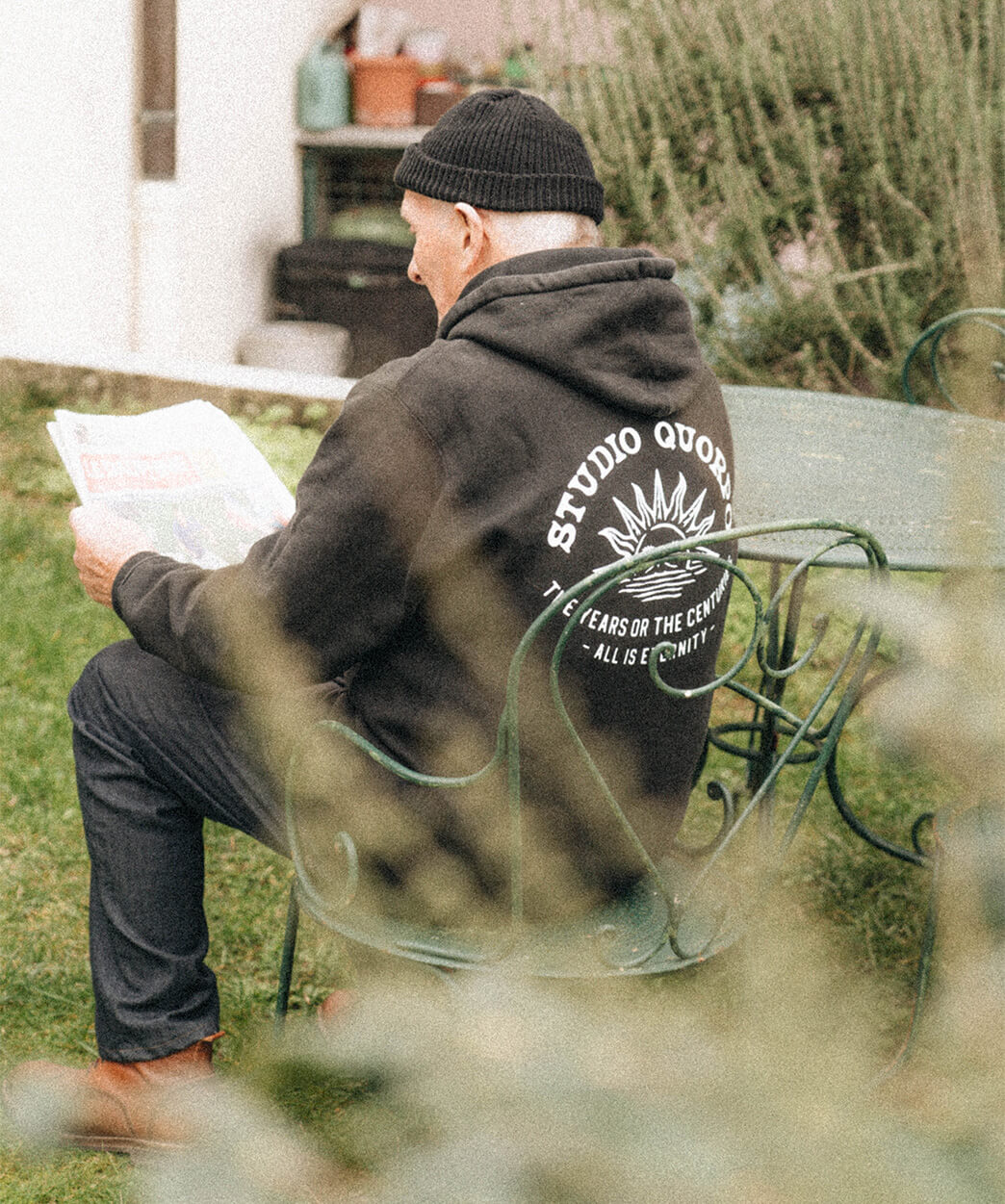 Homme âgé portant un hoodie Studio Quorpo et un bonnet noir, assis à l'extérieur sur une chaise de jardin, lisant un journal.