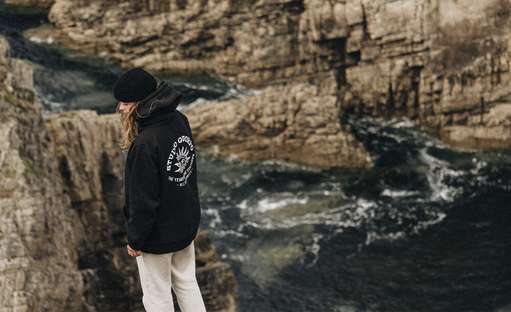 Personne en hoodie Studio Quorpo au bord d'une falaise, face à une mer agitée, symbolisant l'exploration et la liberté en pleine nature.