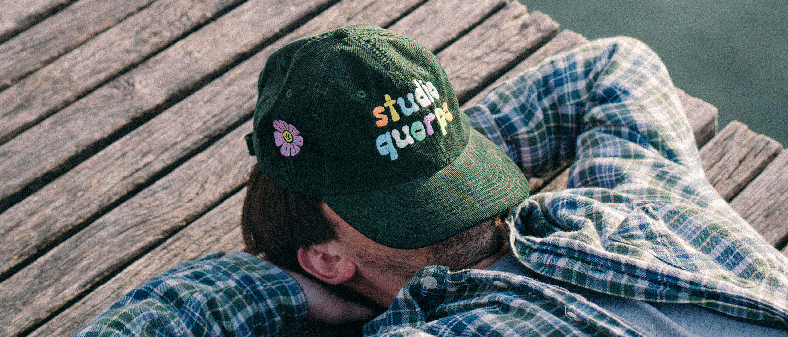 Modèle allongé sur un pont en bois, portant une casquette verte brodée en corduroy avec une inscription colorée Studio Quorpo et une fleur souriante et une chemise à carreaux.